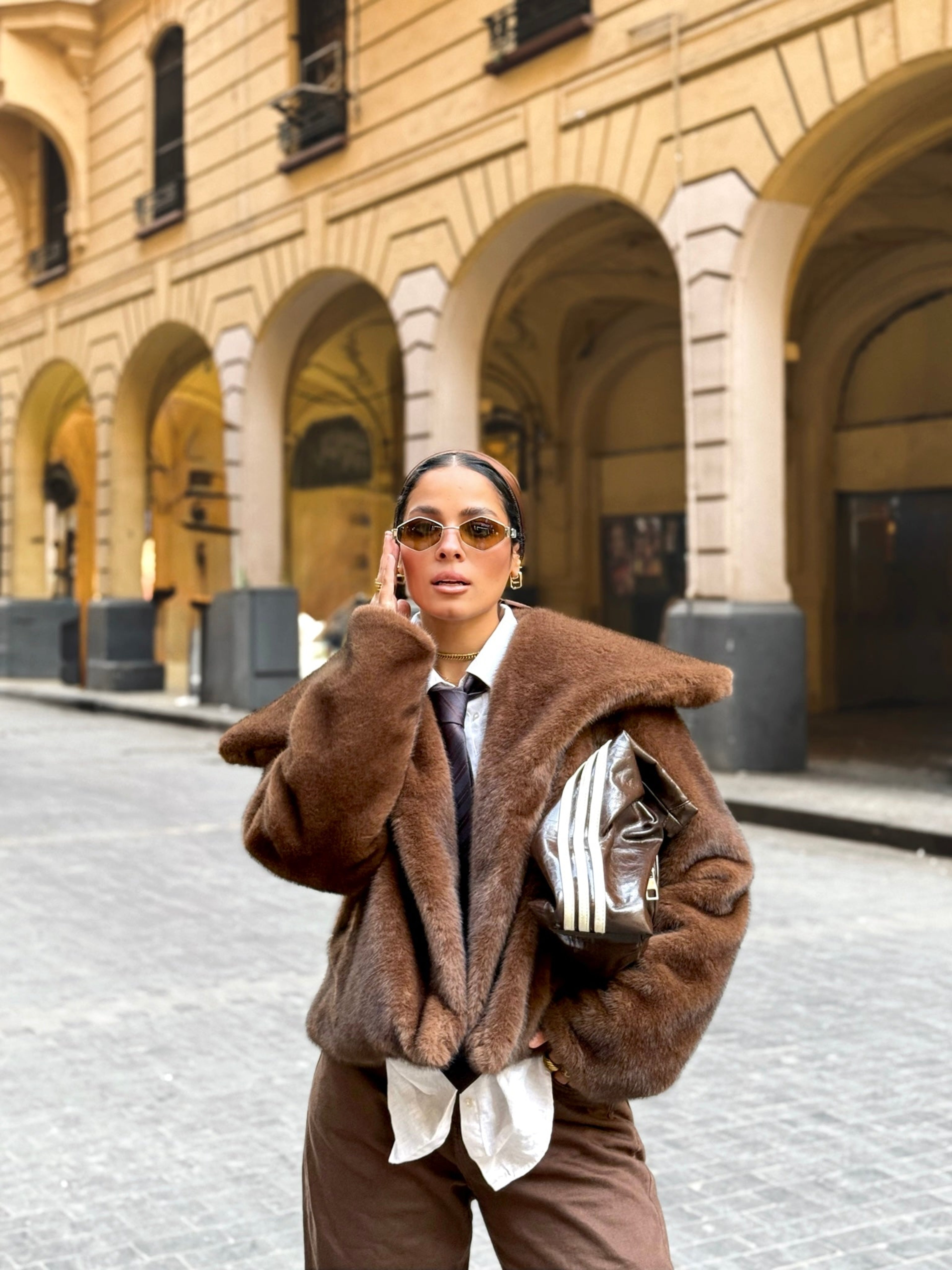 Woman in a brown coat standing in an urban setting with classical architecture.